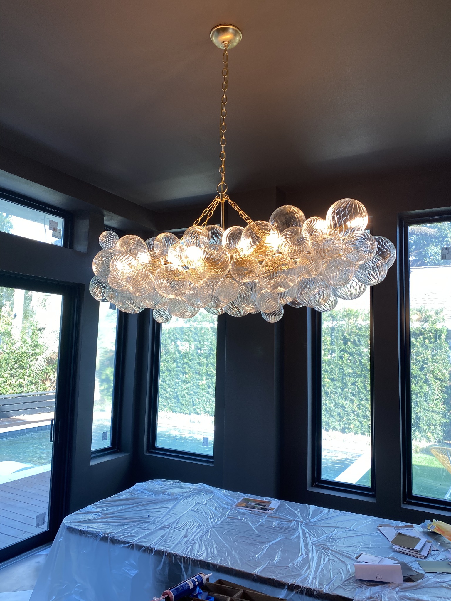 Large clustered glass bubble chandelier newly hung from the ceiling of a dark painted dining room surrounded by windows.