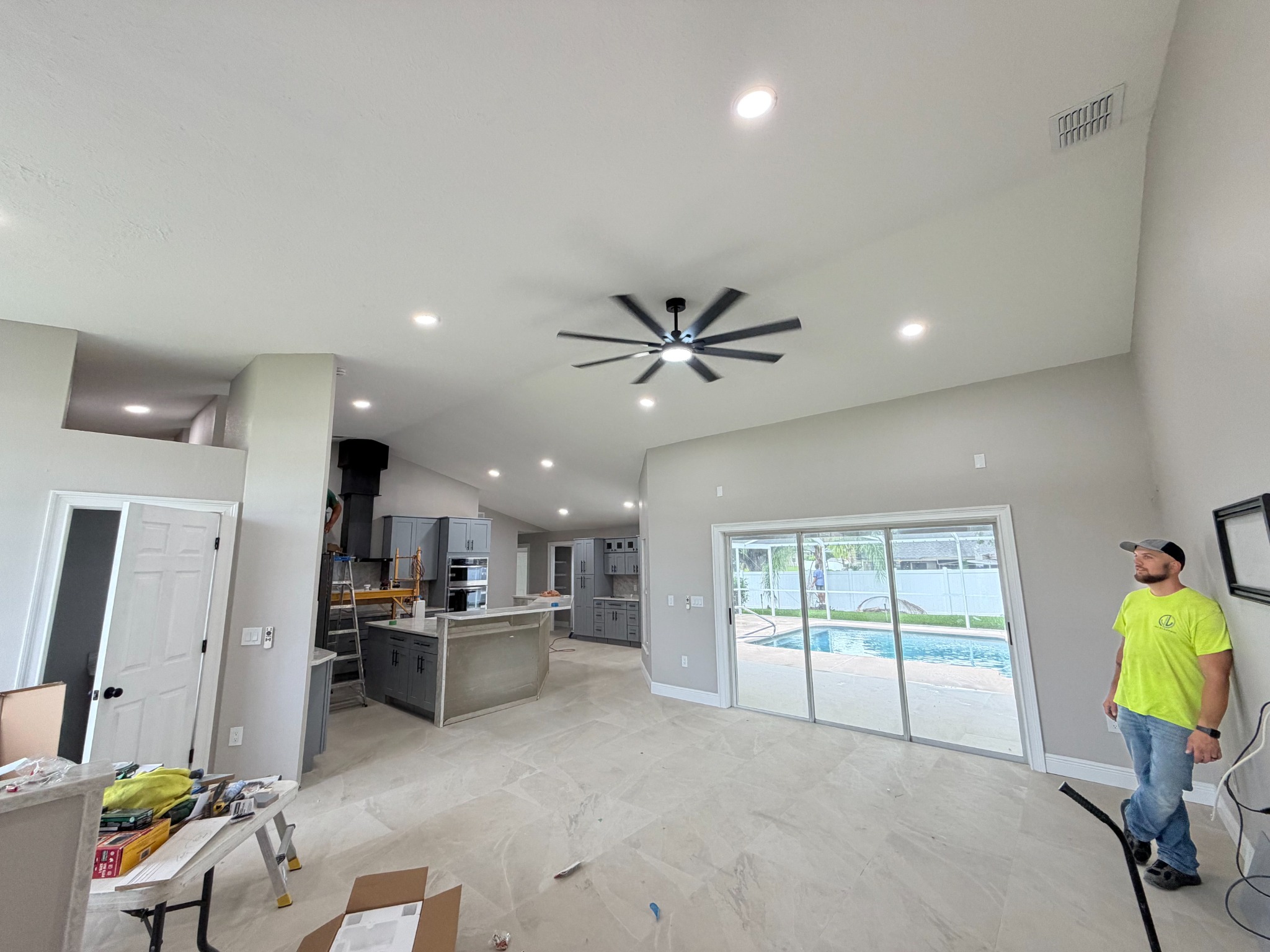 Rooted Electric technician standing in a finished great room lit by recessed LED downlights, with a large ceiling fan and sliding glass doors overlooking a pool.