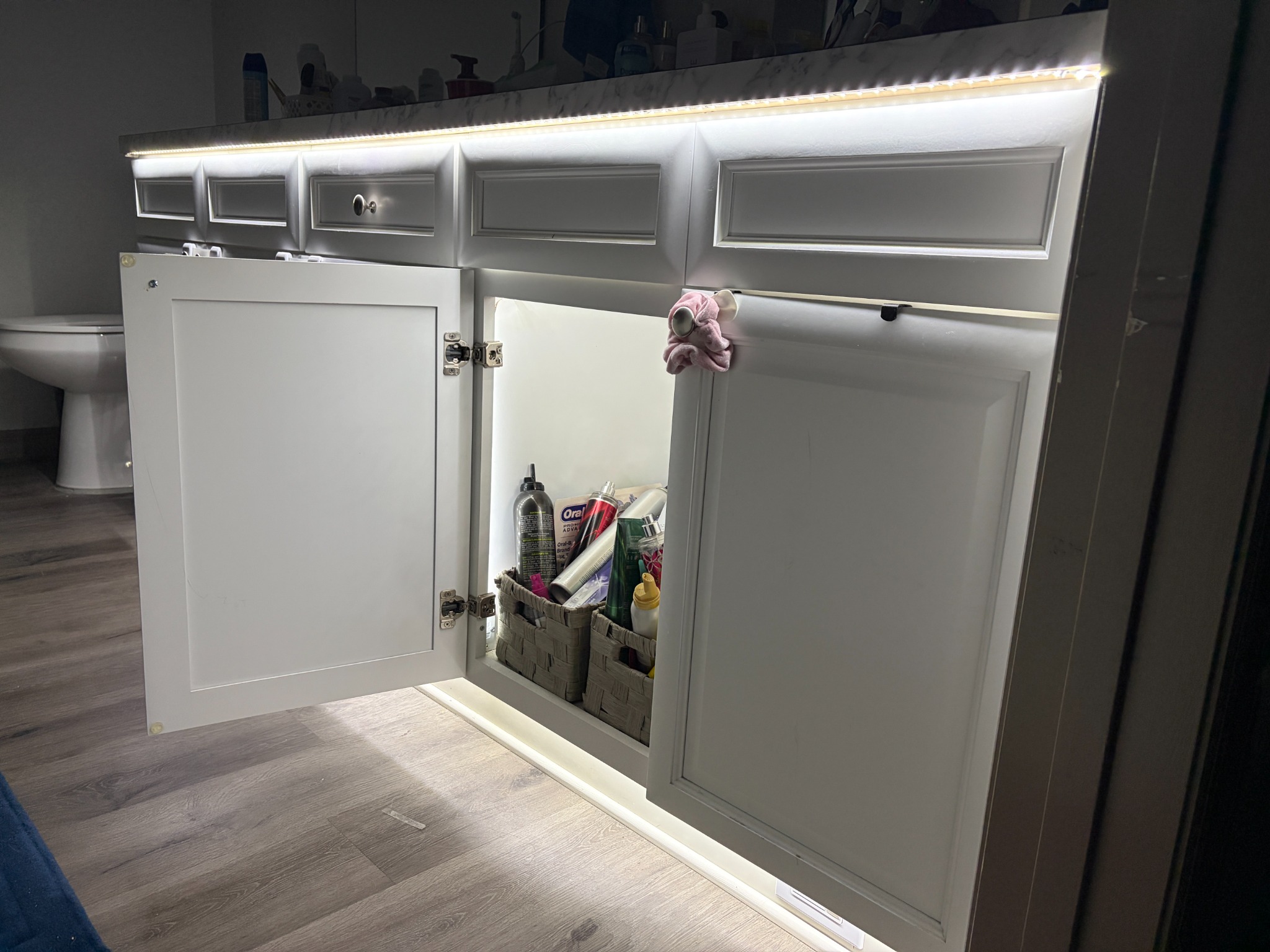 Angled view of a white vanity with a cabinet door open, showing warm white LED strip lighting mounted under the countertop and at the base.