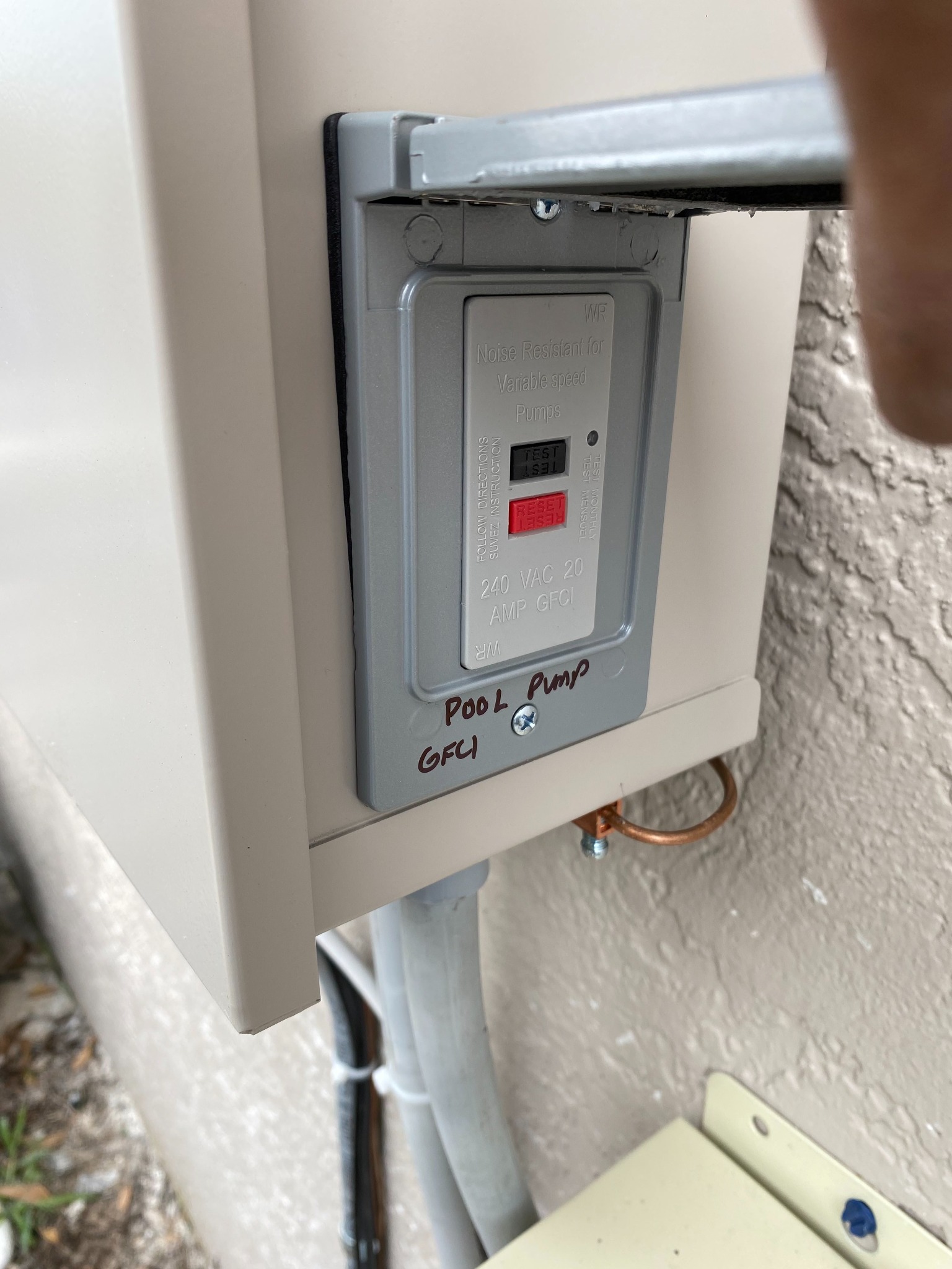 Gray weather-resistant 240 VAC 20 amp GFCI receptacle rated for variable speed pumps, mounted in the side of a beige pool equipment enclosure and hand-labeled Pool Pump GFCI, with a copper bonding lug and conduit below.