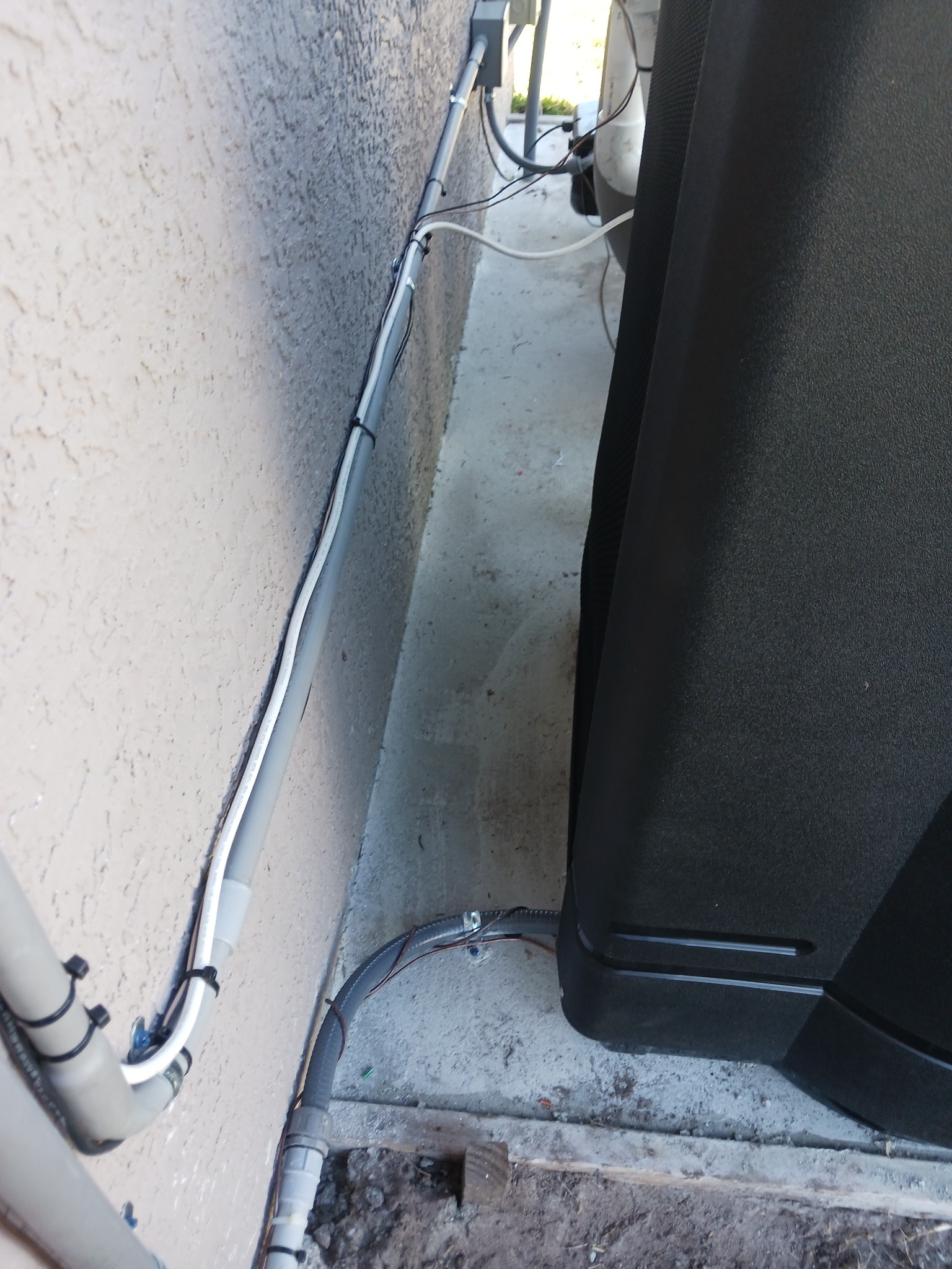 Narrow gap between a stucco house wall and a large black pool equipment cabinet, with gray PVC conduit and white liquid-tight flexible conduit strapped along the wall, cable-tied cable runs, and a weatherproof box feeding the equipment.