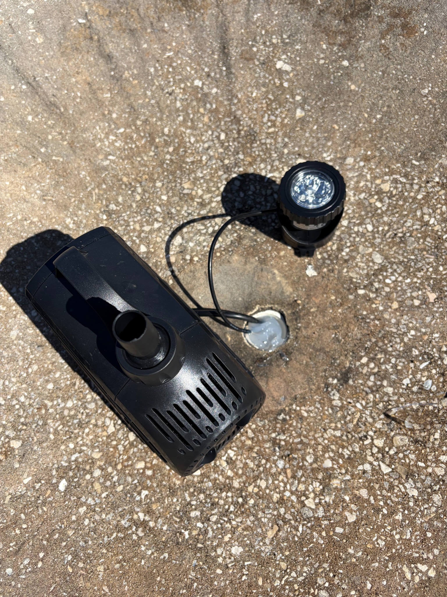 Black submersible fountain pump with a discharge fitting and a small round multi-LED fountain light with its cord, set on a concrete fountain base beside a freshly sealed cord penetration.