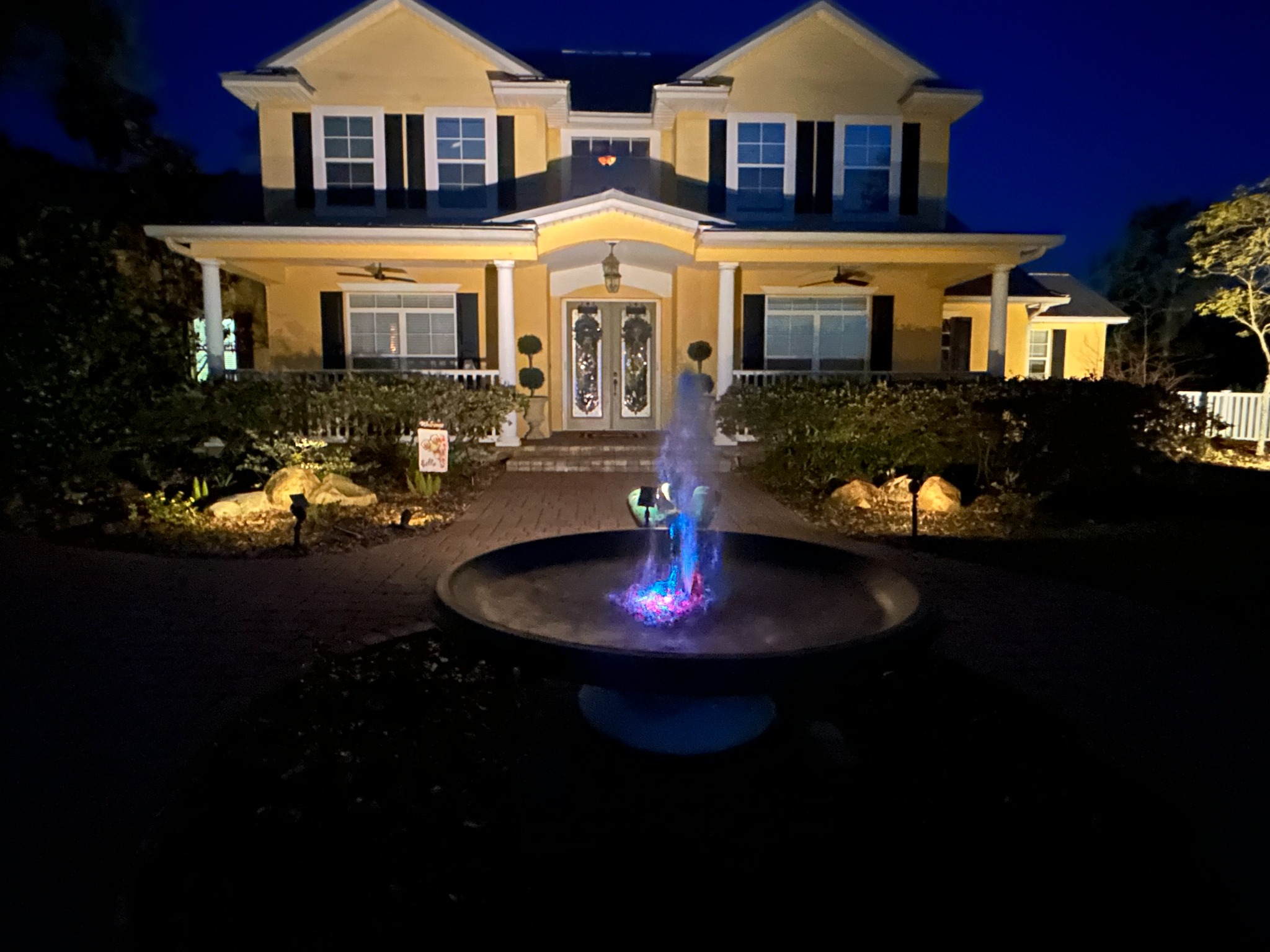 Circular front-yard fountain at night with its spray lit blue and pink from below, path lights along the paver walkway, and warm uplighting washing the two-story home behind it.