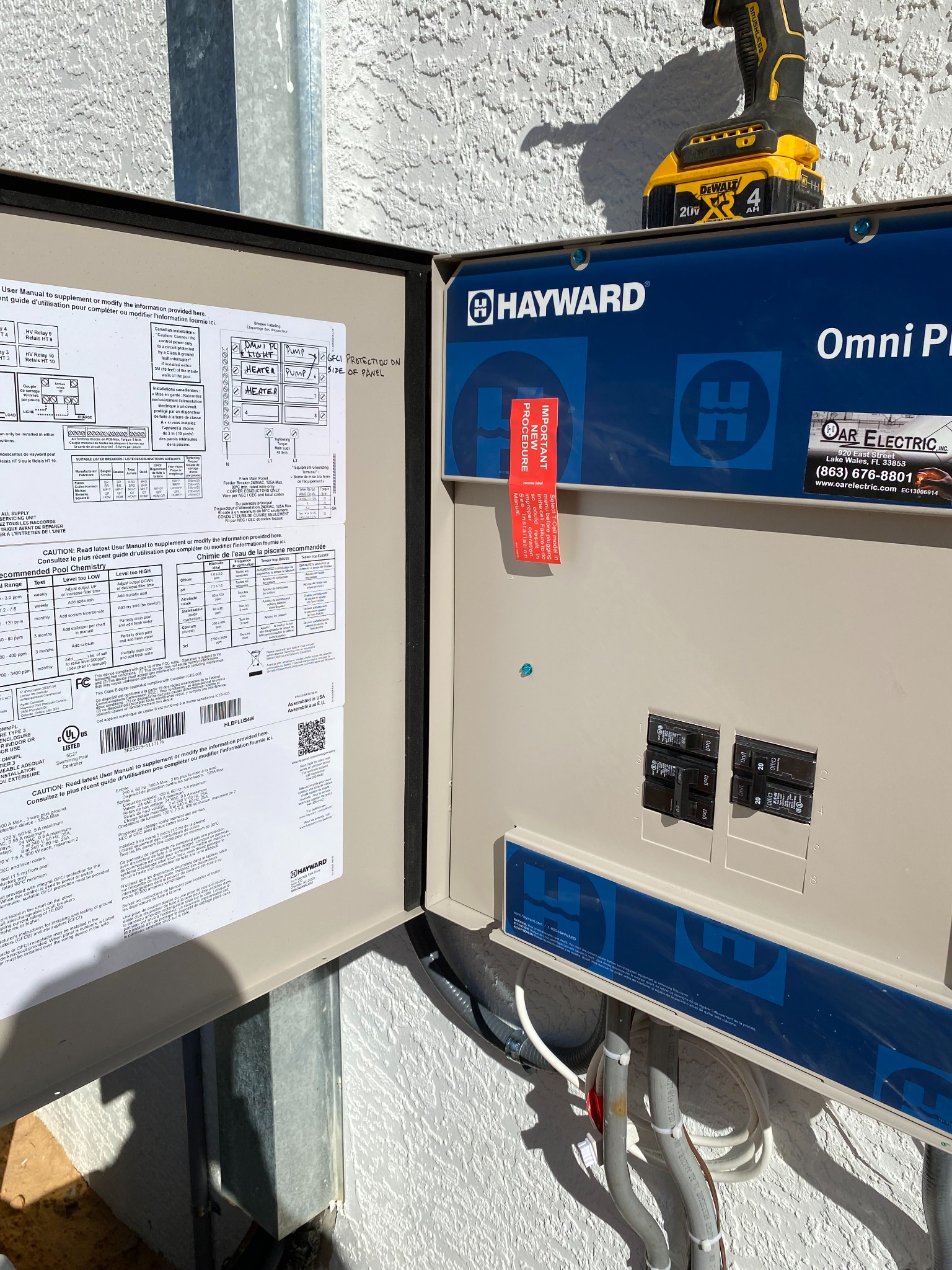 Open Hayward Omni Plus pool control panel in sunlight, with the inside door label sheet hand-marked for the pool light, pump, and heater circuits and a note that GFCI protection is on the side of the panel, and a cordless drill resting on top.
