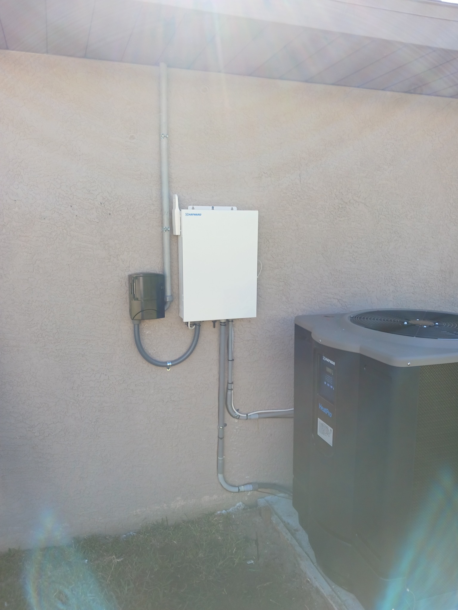 White Hayward OmniLogic automation enclosure mounted on a stucco wall with its wireless antenna alongside, a weatherproof outlet cover fed by liquid-tight flexible conduit, gray PVC conduit running up the wall and down to grade, and a Hayward HeatPro heat pump to the right.
