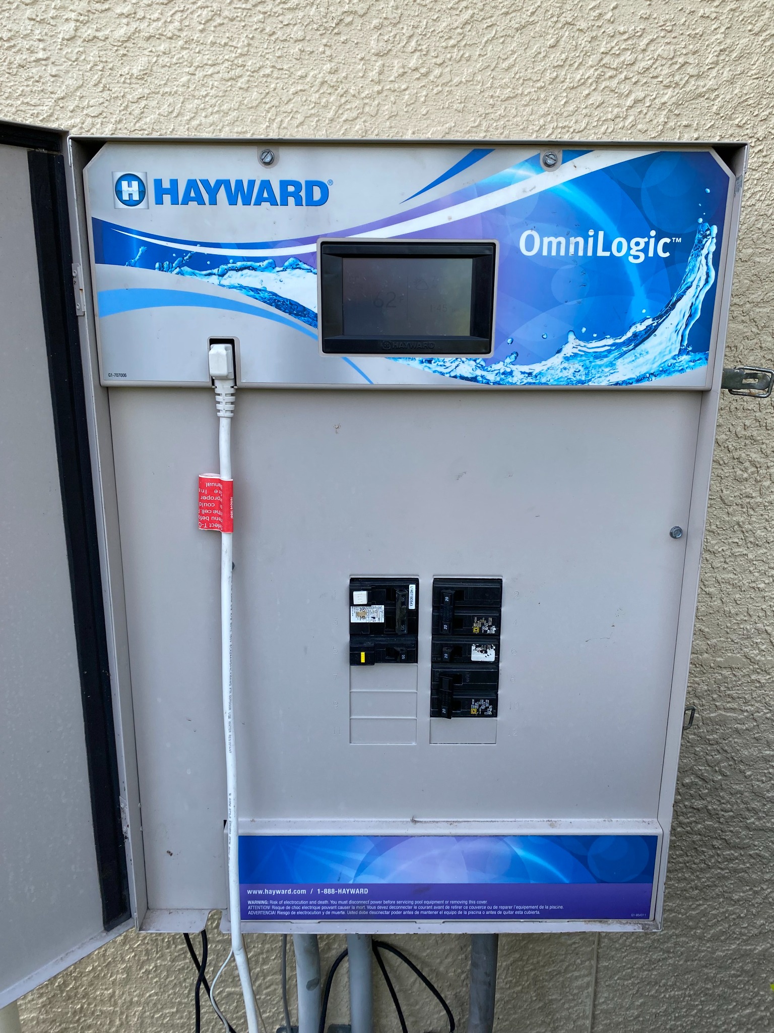 Open Hayward OmniLogic pool automation panel on a stucco wall, showing its touchscreen display and a row of twenty amp breakers in the load center section below.