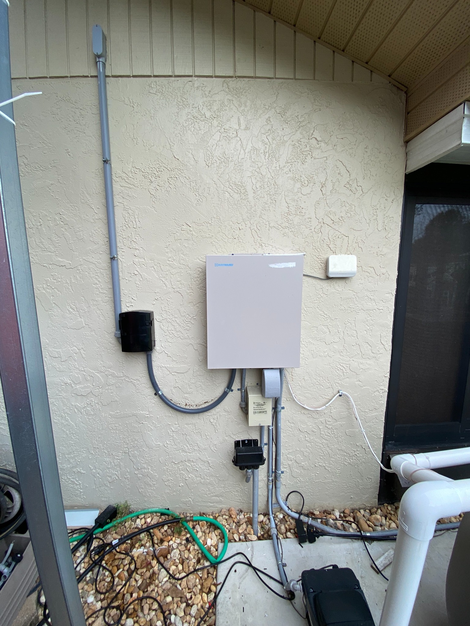 Hayward pool control panel mounted on a stucco wall in a narrow screened courtyard with a wireless antenna beside it, a weatherproof outlet box and transformer below, and conduit dropping into a river rock bed toward the pump.