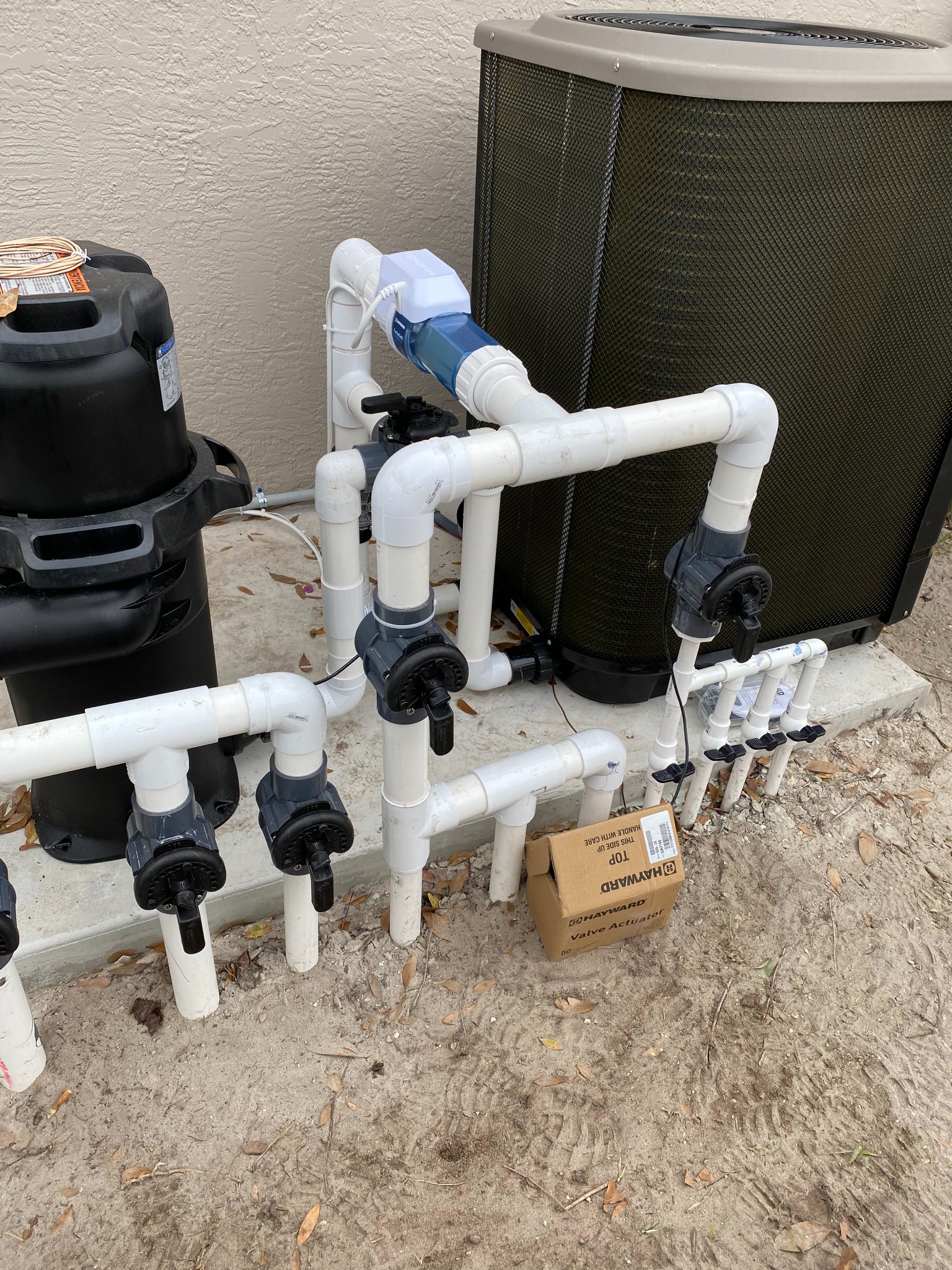 Pool equipment set with a black cartridge filter and a white PVC valve manifold fitted with Hayward automated valve actuators and three-way valves, an inline salt cell, and a heat pump, with a spare valve actuator box on the ground.