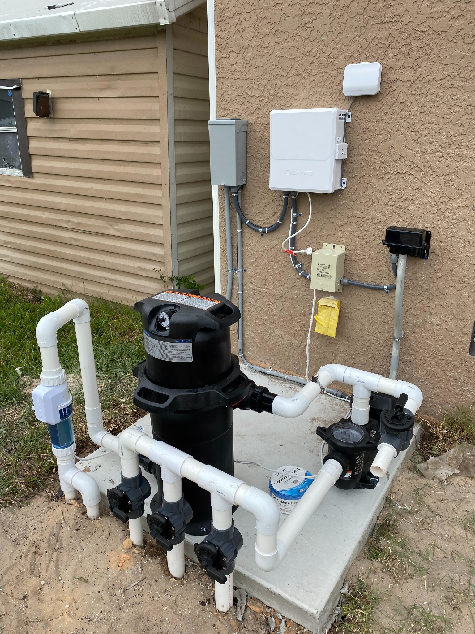 White Hayward pool control box with a wireless antenna mounted above it on a stucco wall, feeding a low-voltage transformer and a weatherproof outlet on a conduit post, with a SwimClear filter, valve manifold, salt cell, and pump on the concrete pad below.