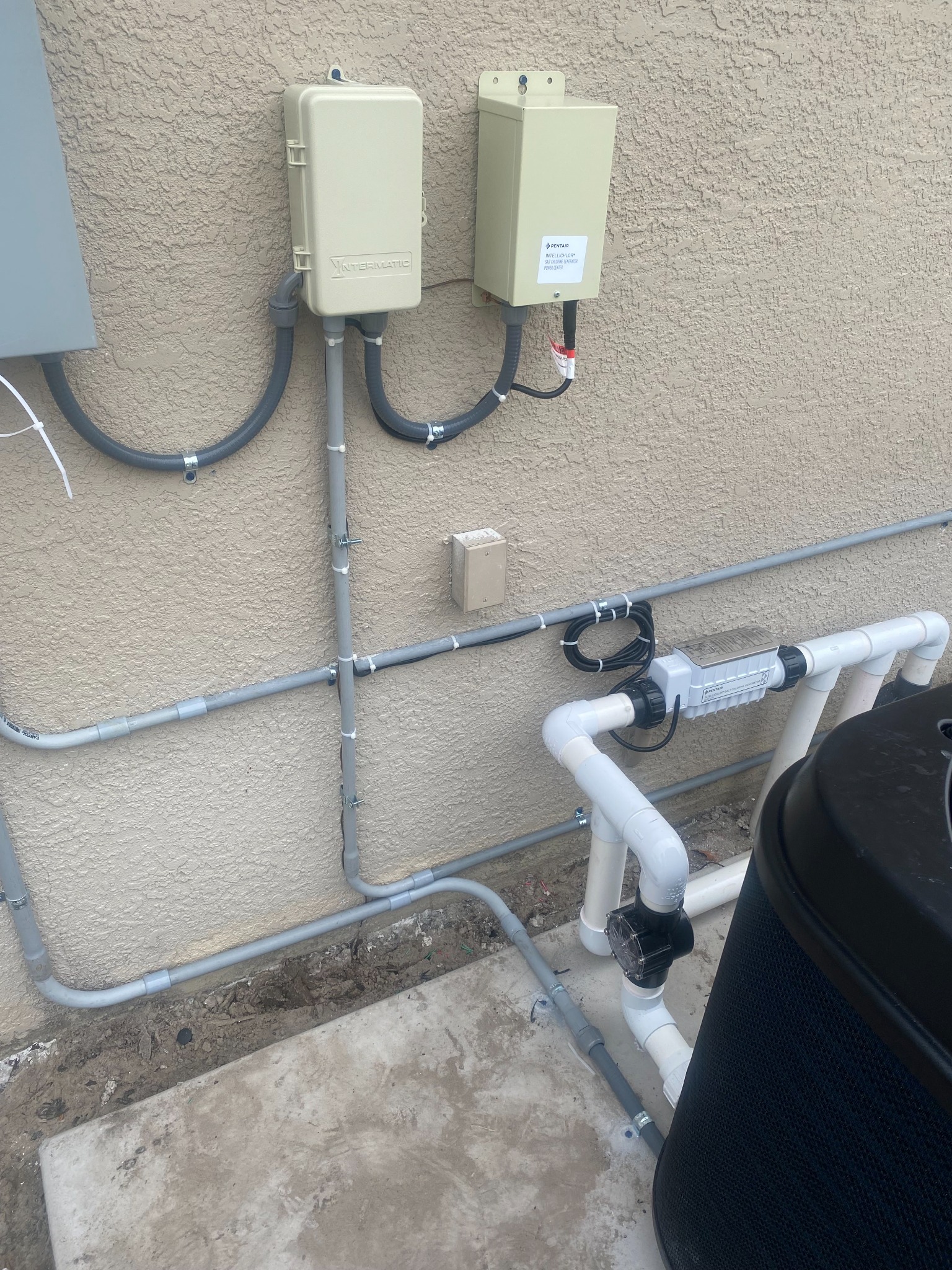 Beige Intermatic timer enclosure and a Pentair IntelliChlor salt chlorine generator power center mounted on a stucco wall, with conduit runs feeding down to a Pentair salt cell plumbed inline in white PVC beside a black pool filter.