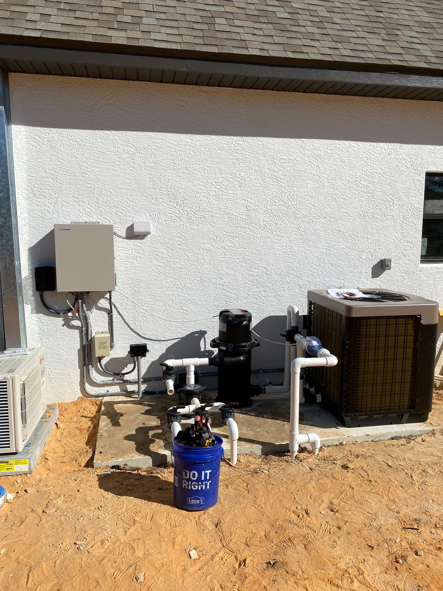 New construction pool equipment pad on bare sand beside a white stucco wall, with a Hayward automation panel and junction box fed by rigid conduit, a black filter and pump plumbed in PVC, and a heat pump on a concrete slab.