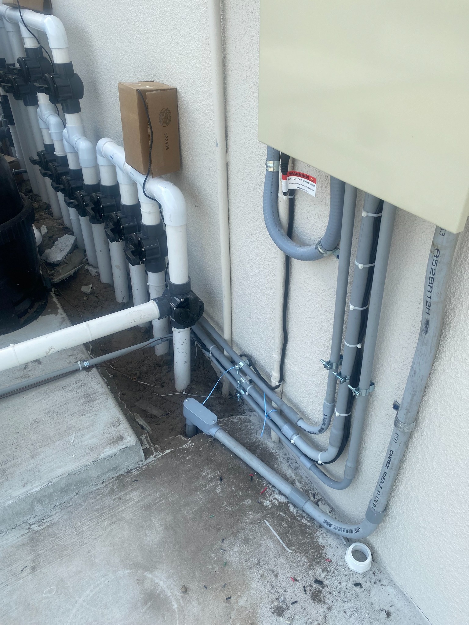 Close view beneath a beige Pentair pool panel showing gray rigid conduits and a liquidtight flexible whip strapped to the stucco wall and turned along the deck toward a white PVC valve manifold.