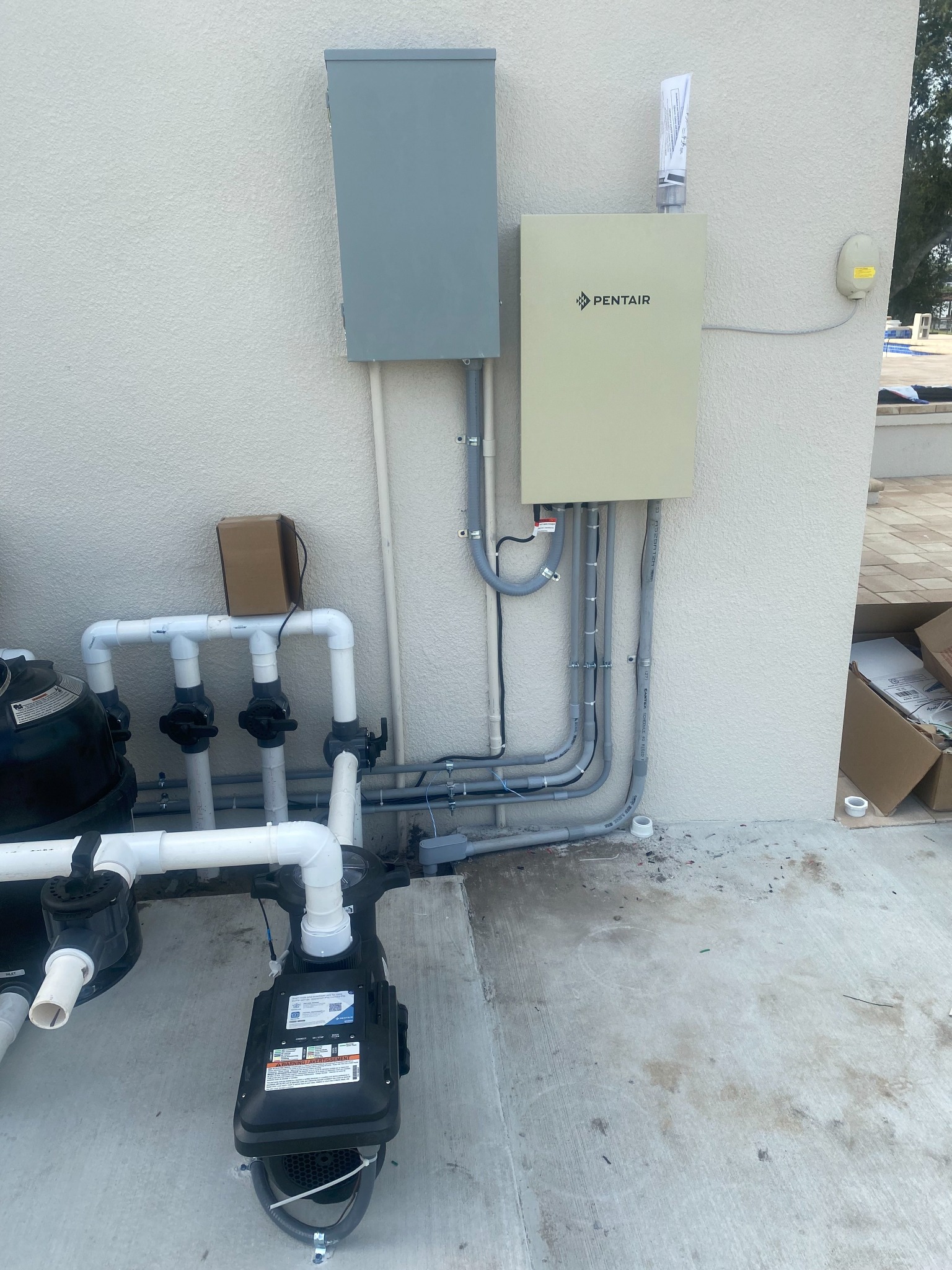 Gray load center and a beige Pentair pool automation panel mounted side by side on a stucco wall, with several gray conduits routed down to a Pentair variable speed pump and a filter valve manifold on the pool deck.