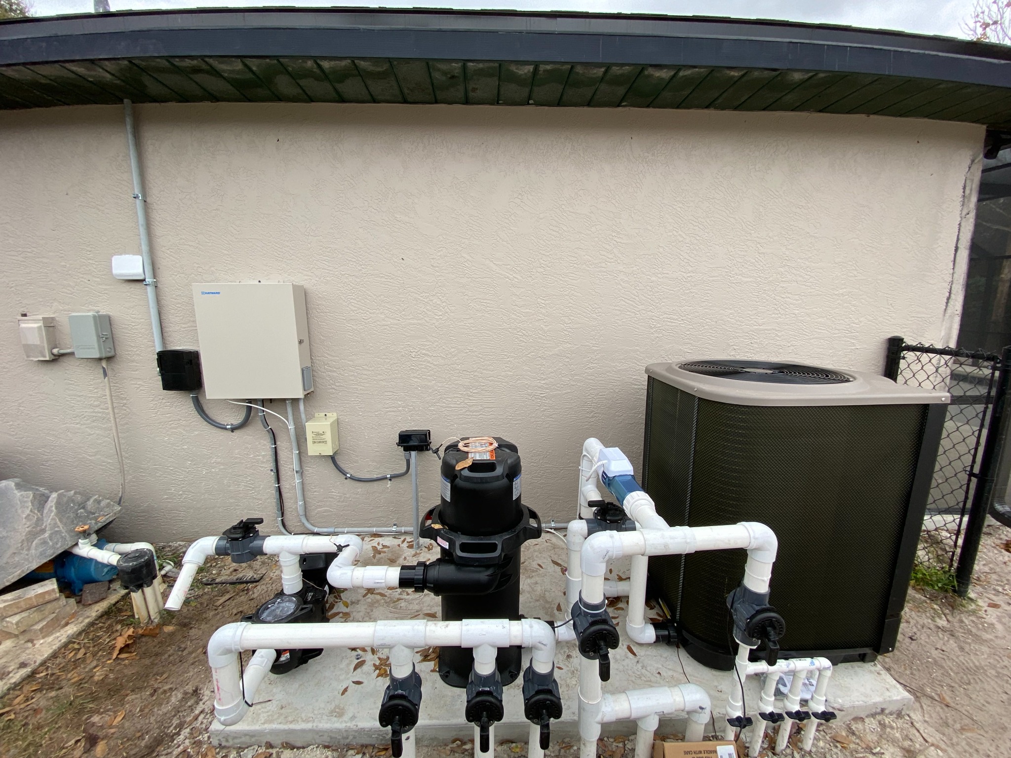 Pool equipment pad against a stucco wall with a Hayward control panel and disconnect boxes fed by rigid conduit, a black cartridge filter and pump plumbed in white PVC, an inline salt chlorinator cell, and a heat pump beside a chain link gate.