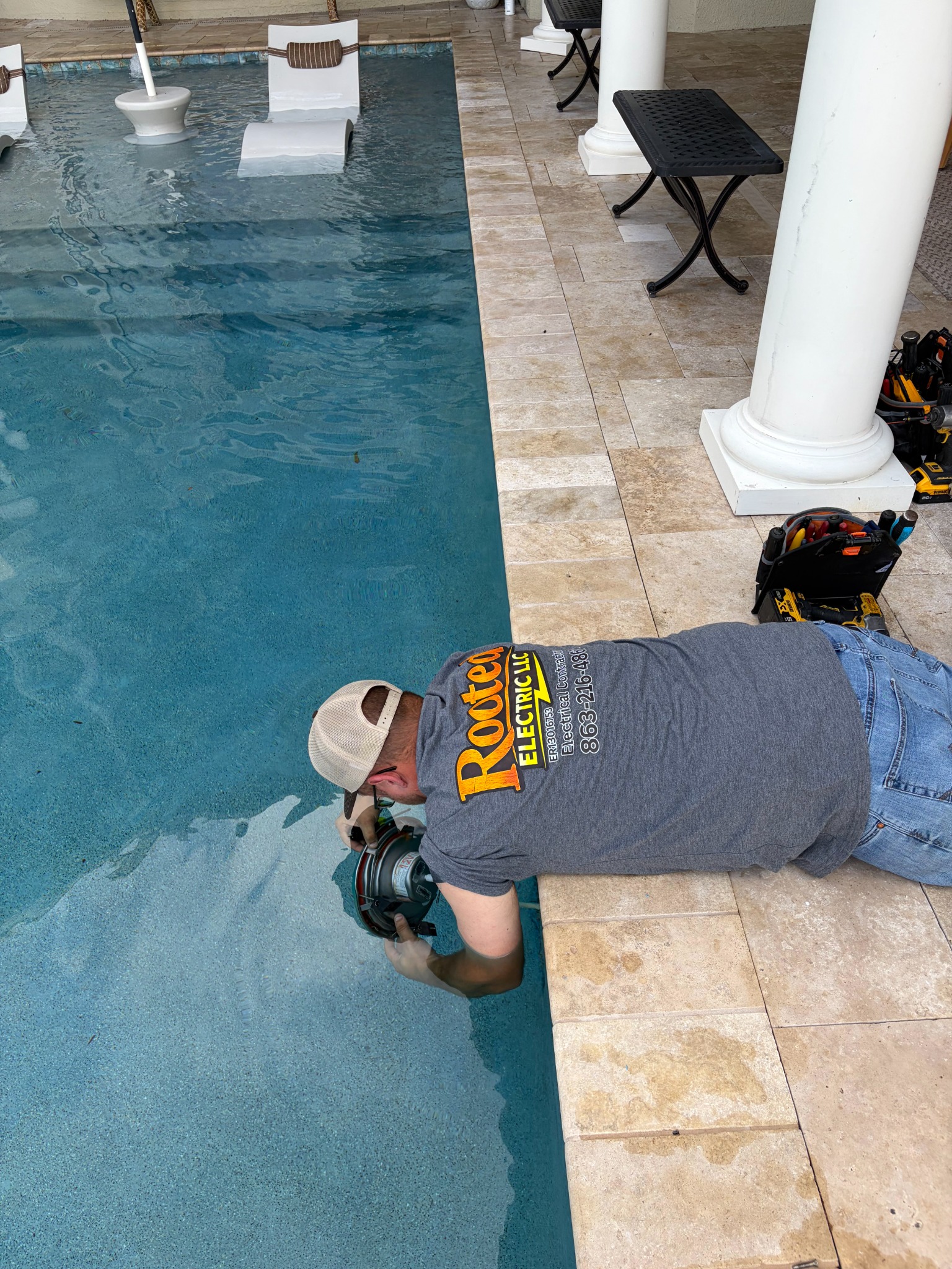 Rooted Electric electrician in a branded shirt lying on a travertine pool deck, reaching into the water to fit a round pool light fixture into its niche, with tool bags and cordless tools staged on the deck behind.