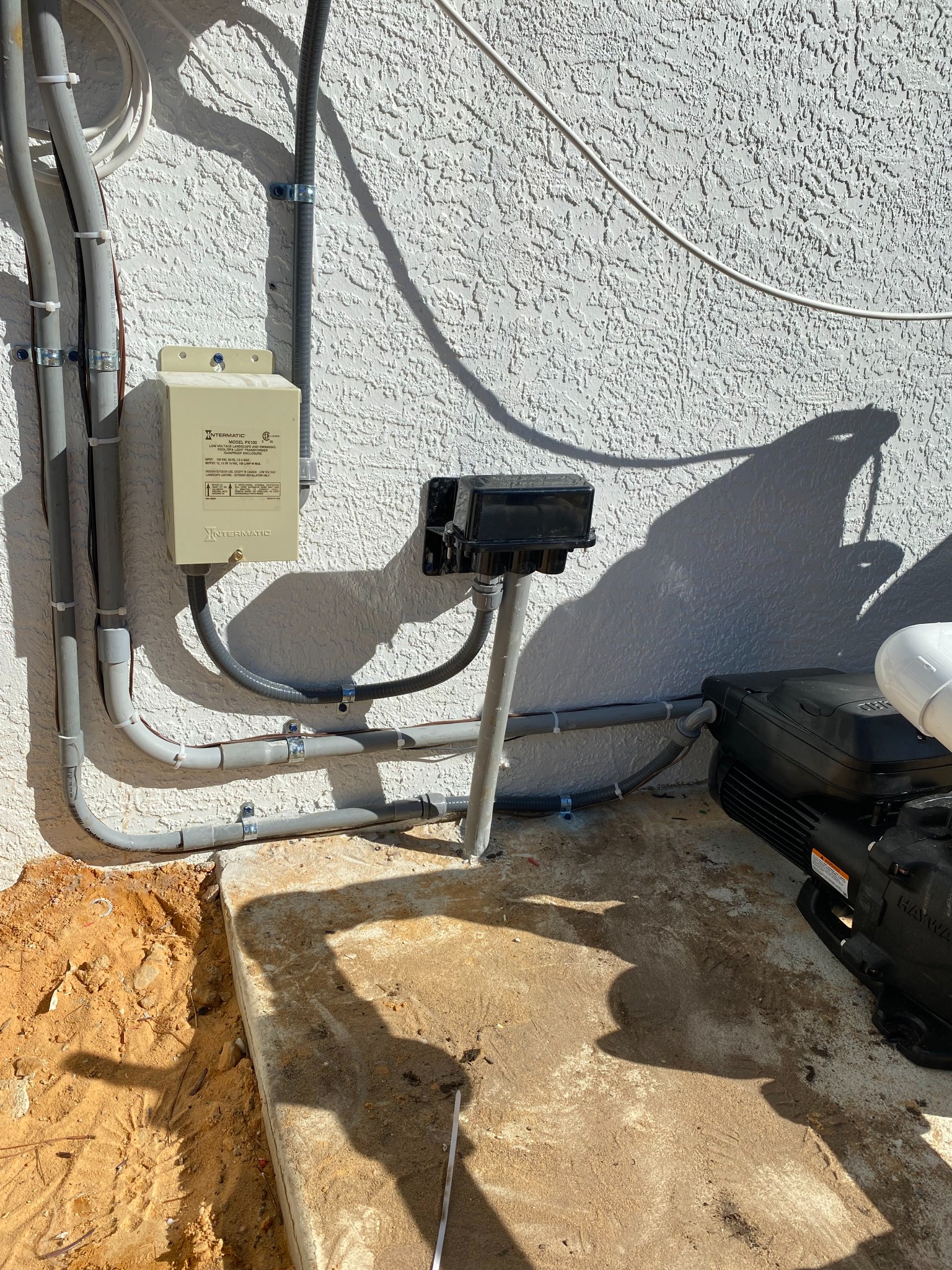 Intermatic PX100 pool light transformer on a white stucco wall with neatly strapped gray conduit runs feeding a black weatherproof receptacle box on a post, and a Hayward pump on the concrete pad at right.