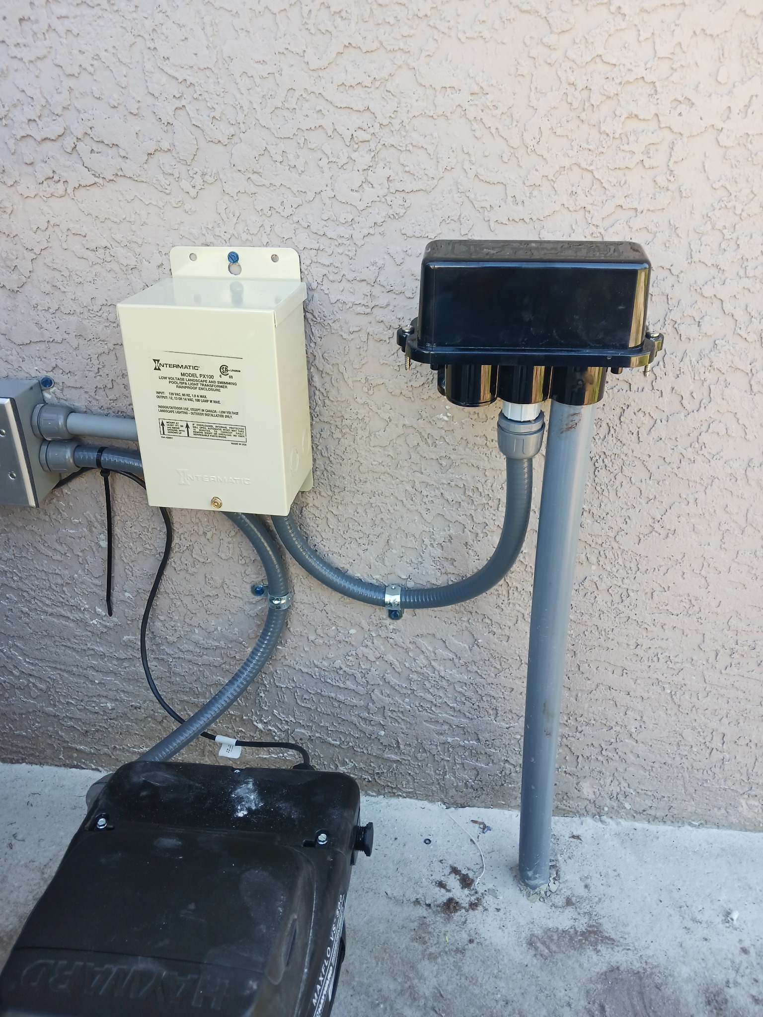 Intermatic PX100 low-voltage landscape and swimming pool light transformer mounted on a stucco wall, connected with liquidtight flexible conduit to a black weatherproof outlet box on a gray rigid conduit post, with a Hayward pump below.