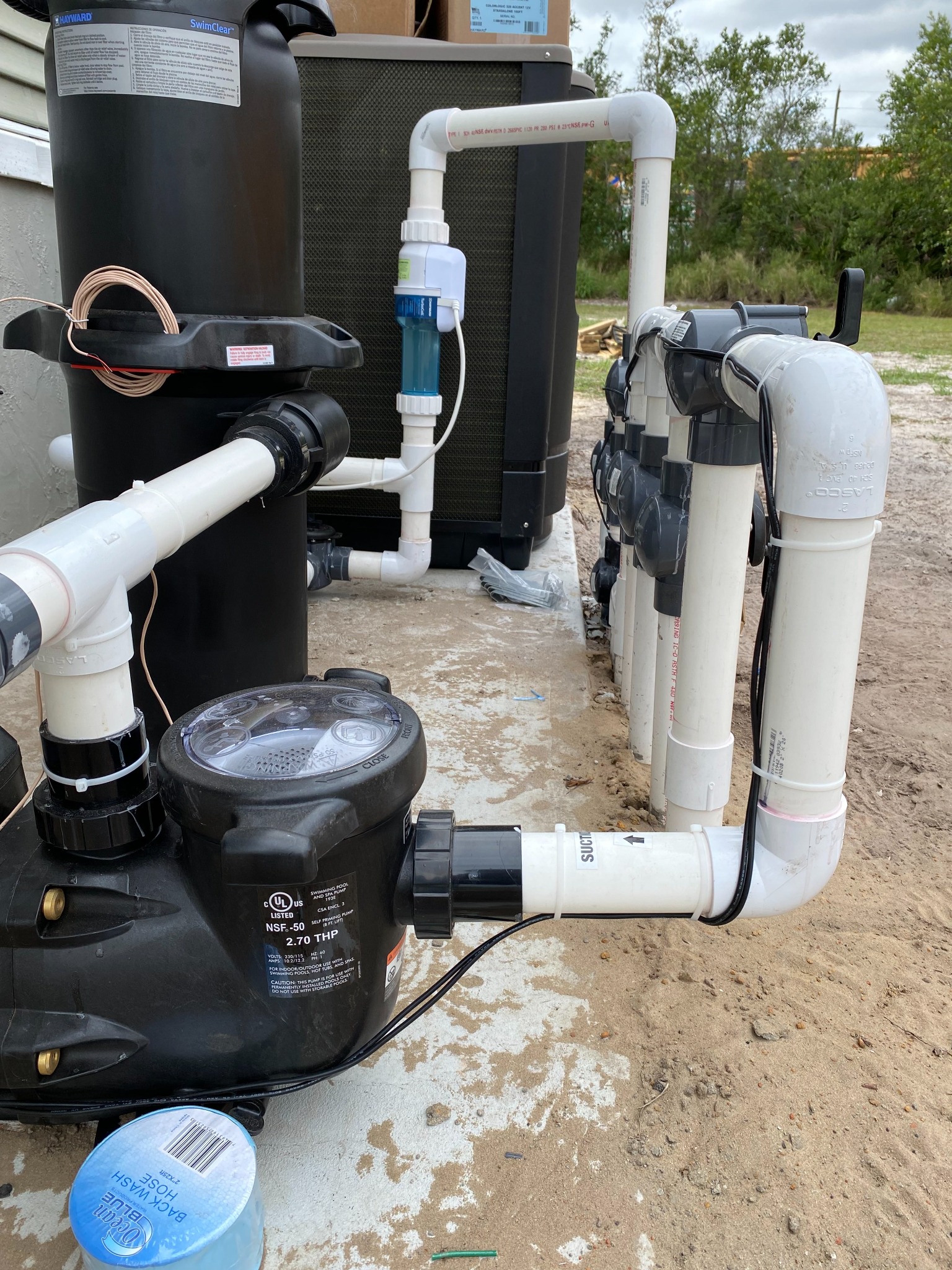 Hayward SwimClear cartridge filter with a coiled bare copper bonding wire on top, a black 2.7 total horsepower pool and spa pump, an inline salt chlorinator cell, and a white PVC valve manifold on a concrete and sand pad.