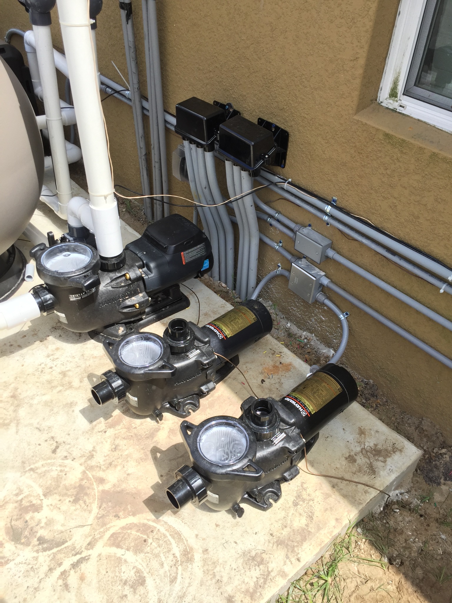 Concrete pool equipment pad holding a Hayward TriStar VS variable-speed pump plumbed to a filter with two more Hayward Max-Flo pumps staged beside it, fed by gray PVC conduits from two black weatherproof enclosures and metal junction boxes strapped to the stucco wall, with bare copper bonding wire run to each pump.