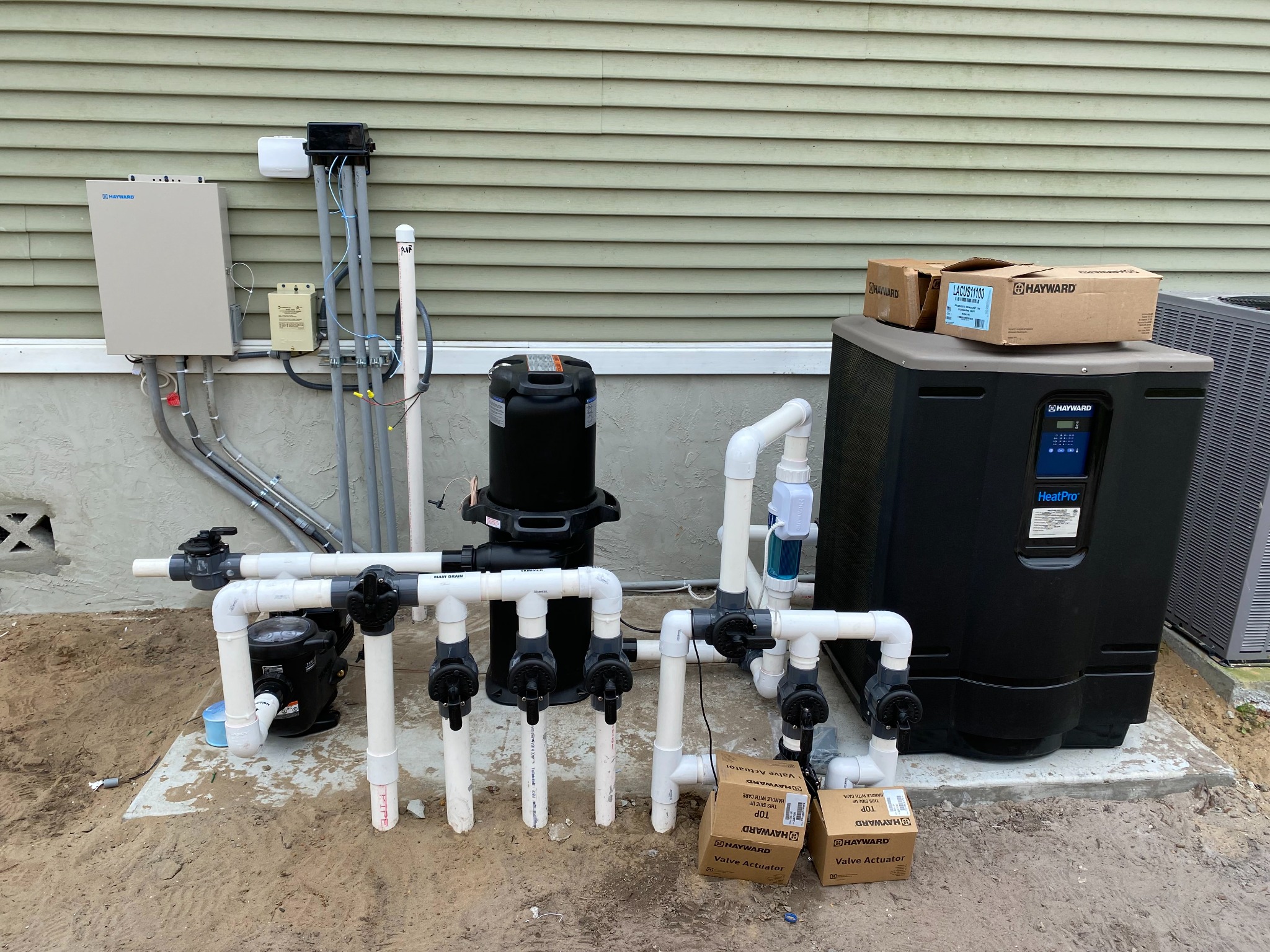 Raised Hayward automation panel, low-voltage transformer, and a black weatherproof pool-light junction box mounted on green siding above the equipment pad, fed by gray conduit risers, with a labeled main drain and skimmer valve manifold, cartridge filter, pump, inline salt cell, and a Hayward HeatPro heat pump on the pad below.