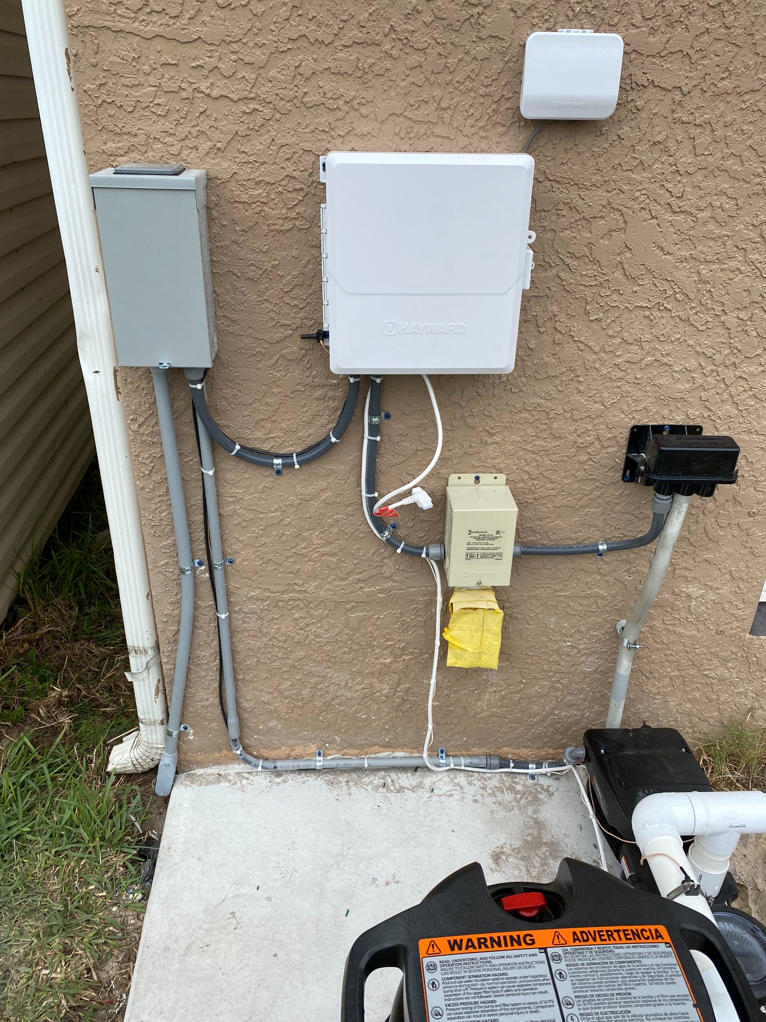 Hayward pool control box on a tan stucco wall with a wireless antenna above it, a gray disconnect at left, an Intermatic transformer feeding a black weatherproof outlet box on a post, and conduit strapped down the wall toward the pump.