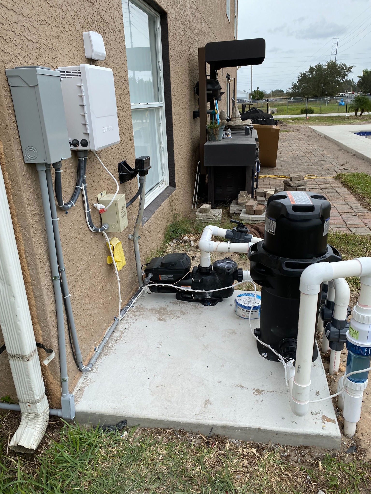 White pool automation control box and a wireless antenna mounted on a tan stucco wall next to a gray disconnect, with conduit and a transformer feeding down to a variable speed pump, cartridge filter, and salt cell on a concrete pad.